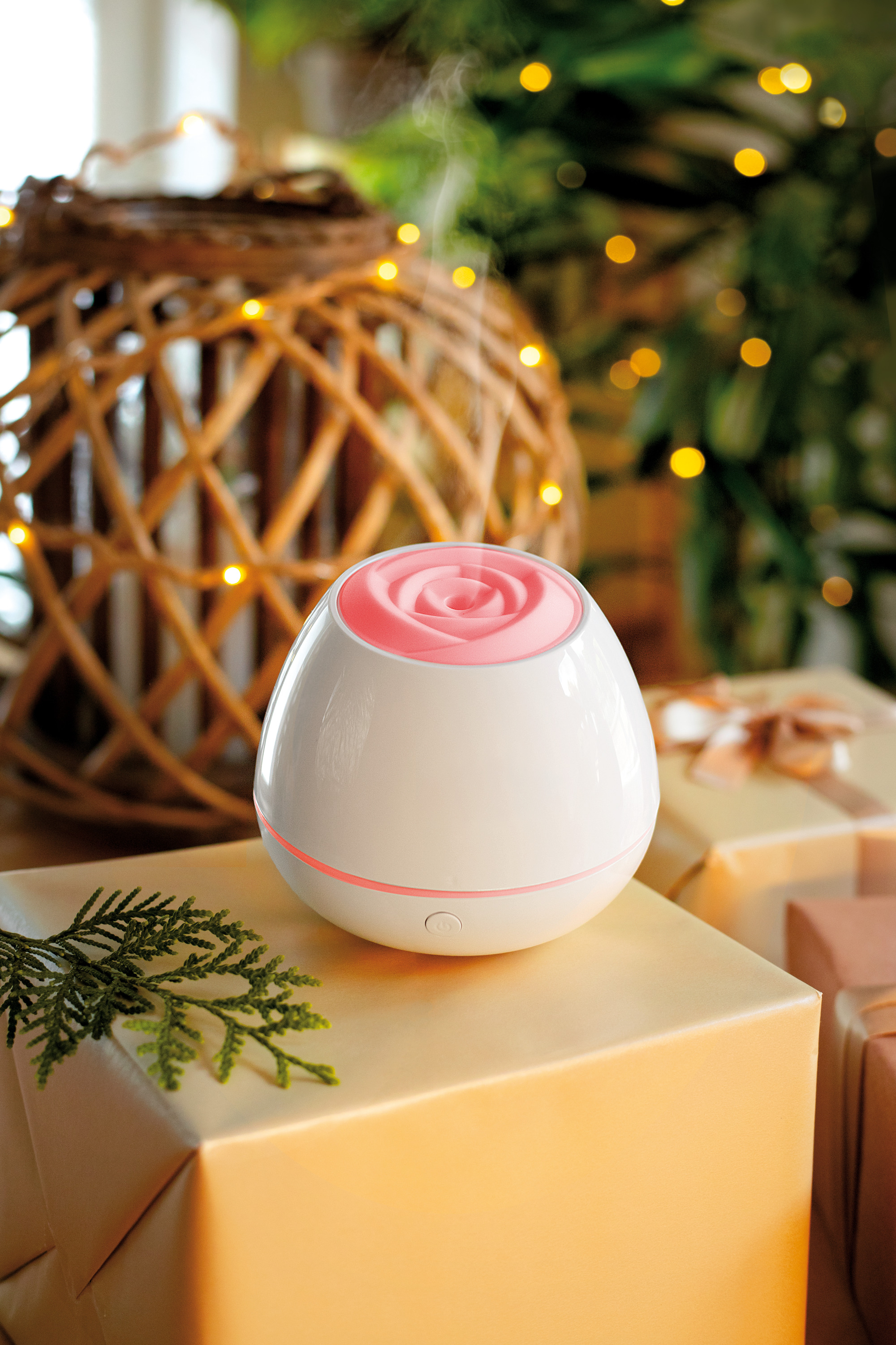 Weißer, eiförmiger Diffusor mit rosa Rosenaufsatz. Steht auf einer Geschenkbox. Hintergrund: Weihnachtsbaum.