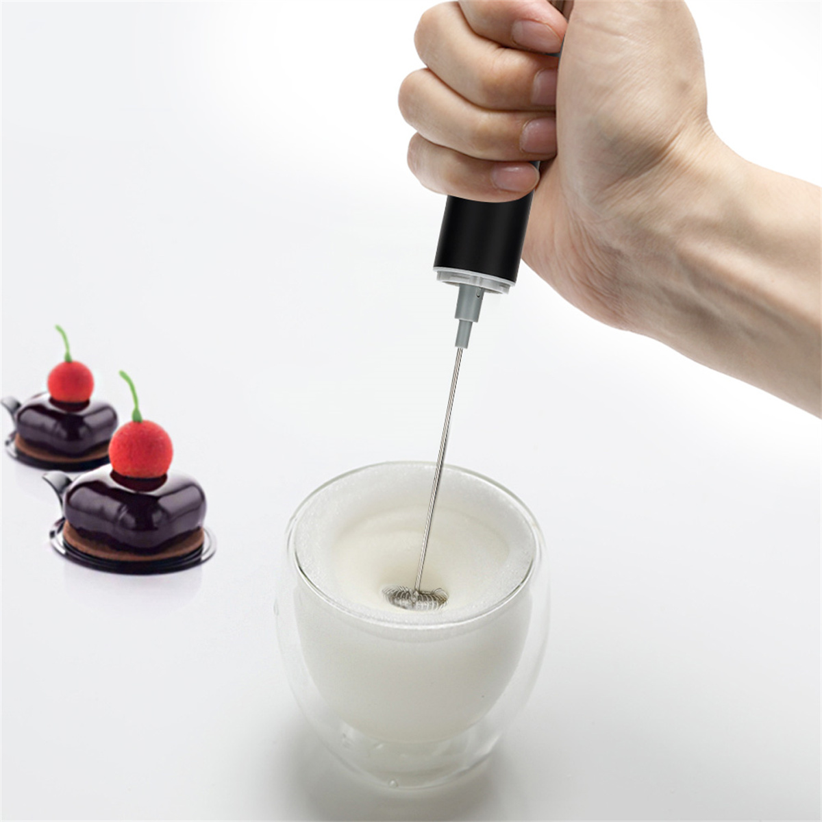 Eine Hand schäumt Milch in einem Glas mit einem Mixer auf. Zwei kleine Kuchen mit Kirschen liegen auf weißem Untergrund.