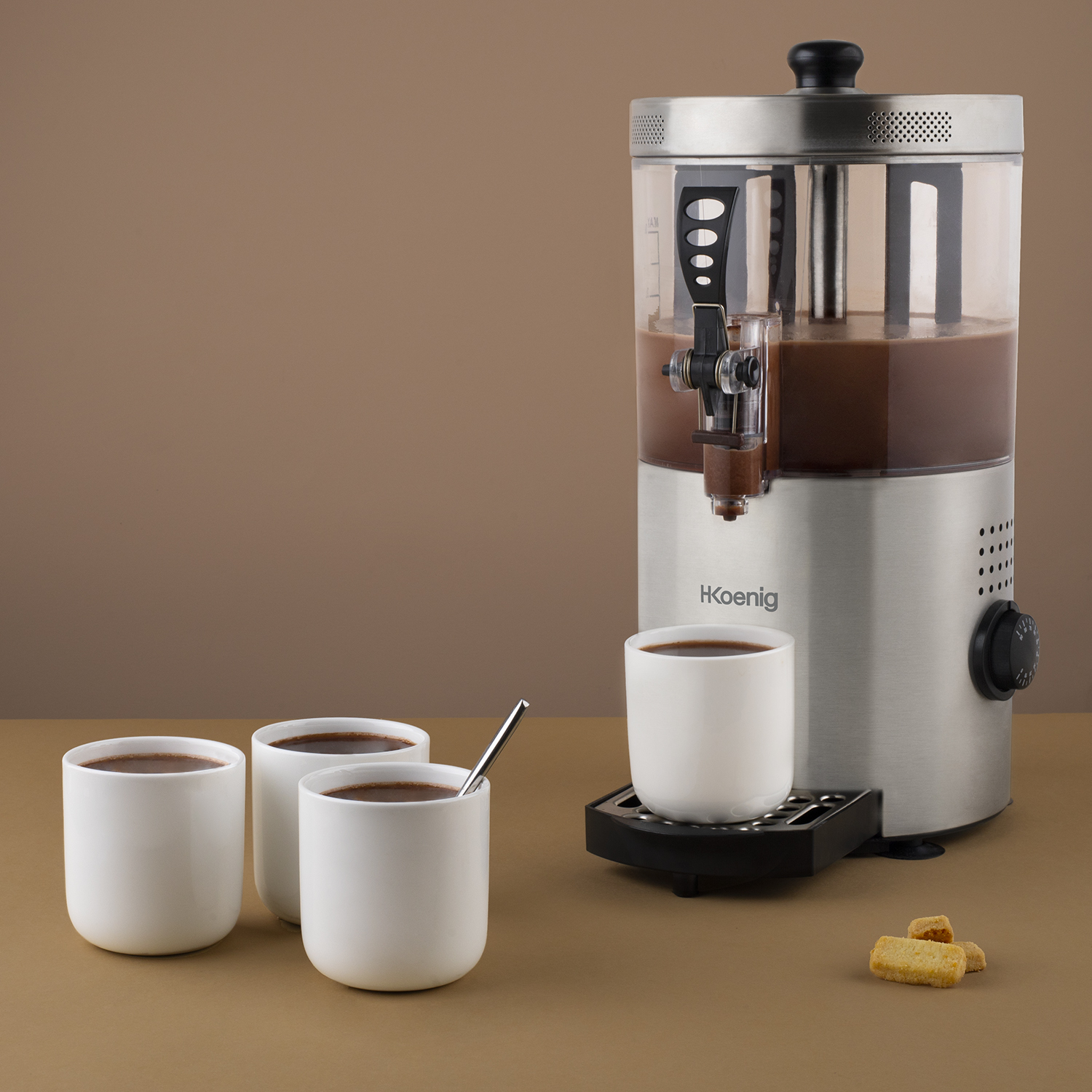 Hot chocolate machine with cups and cookies on a brown surface.