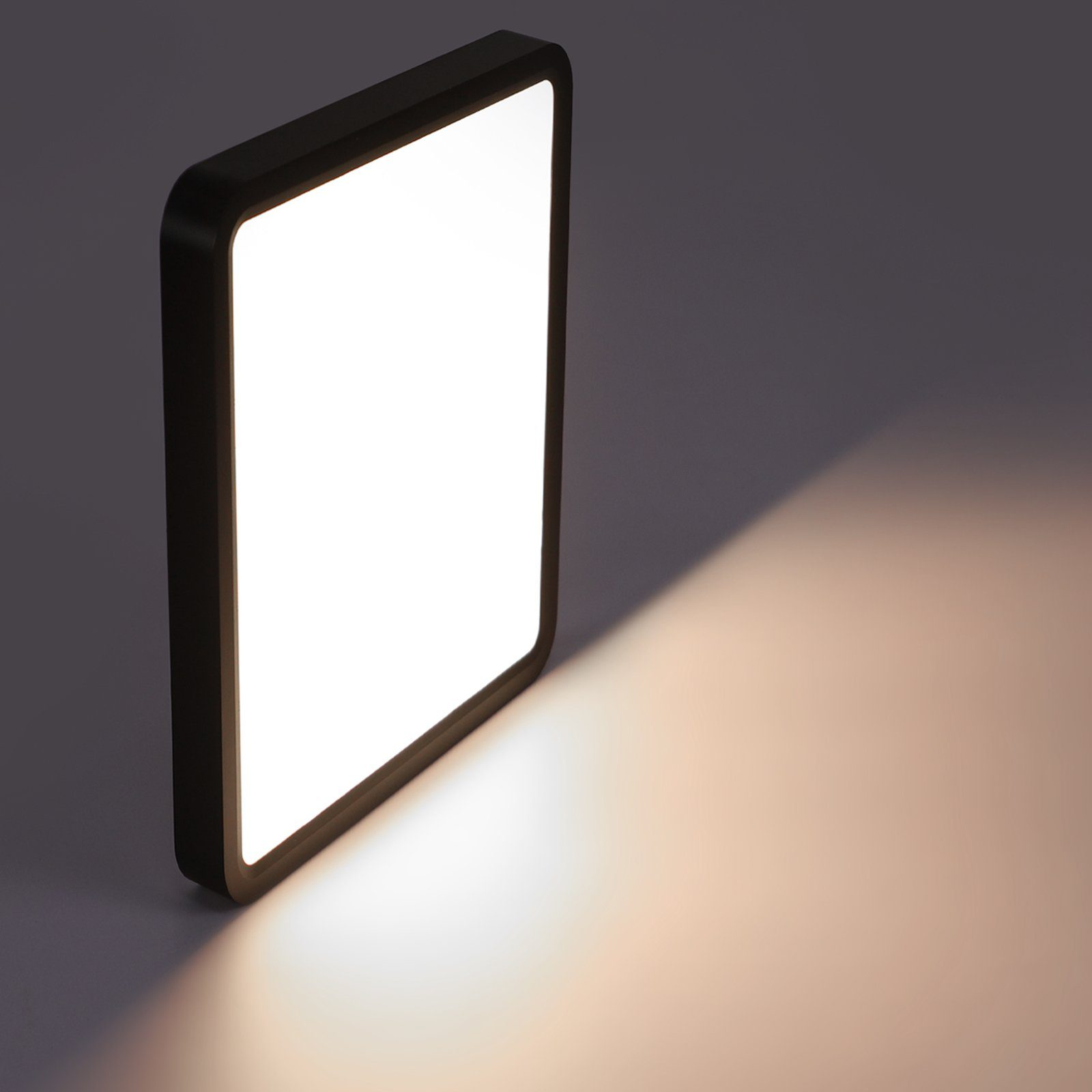 Podświetlony kwadratowy panel z czarną ramą stoi pod kątem, rzucając cień na szarą powierzchnię.