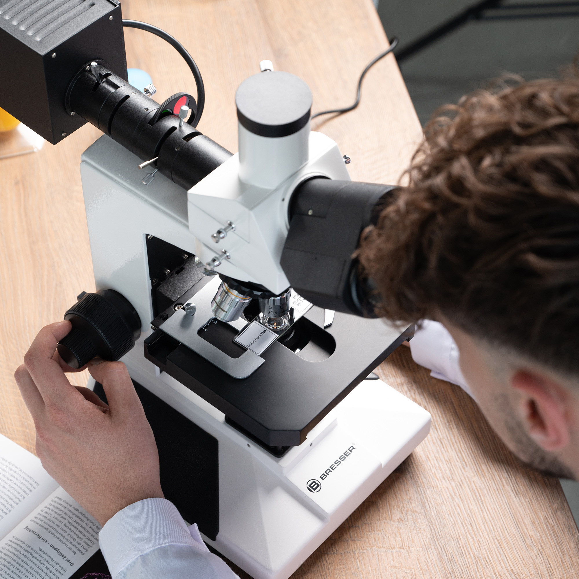 Una persona ajusta un microscopio blanco y negro sobre una mesa de madera, junto a un libro.
