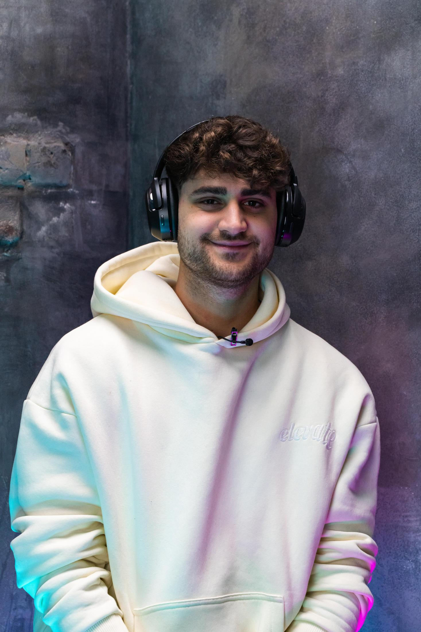 Młody mężczyzna z kręconymi włosami, w białej bluzie z kapturem i czarnych słuchawkach, uśmiechnięty.