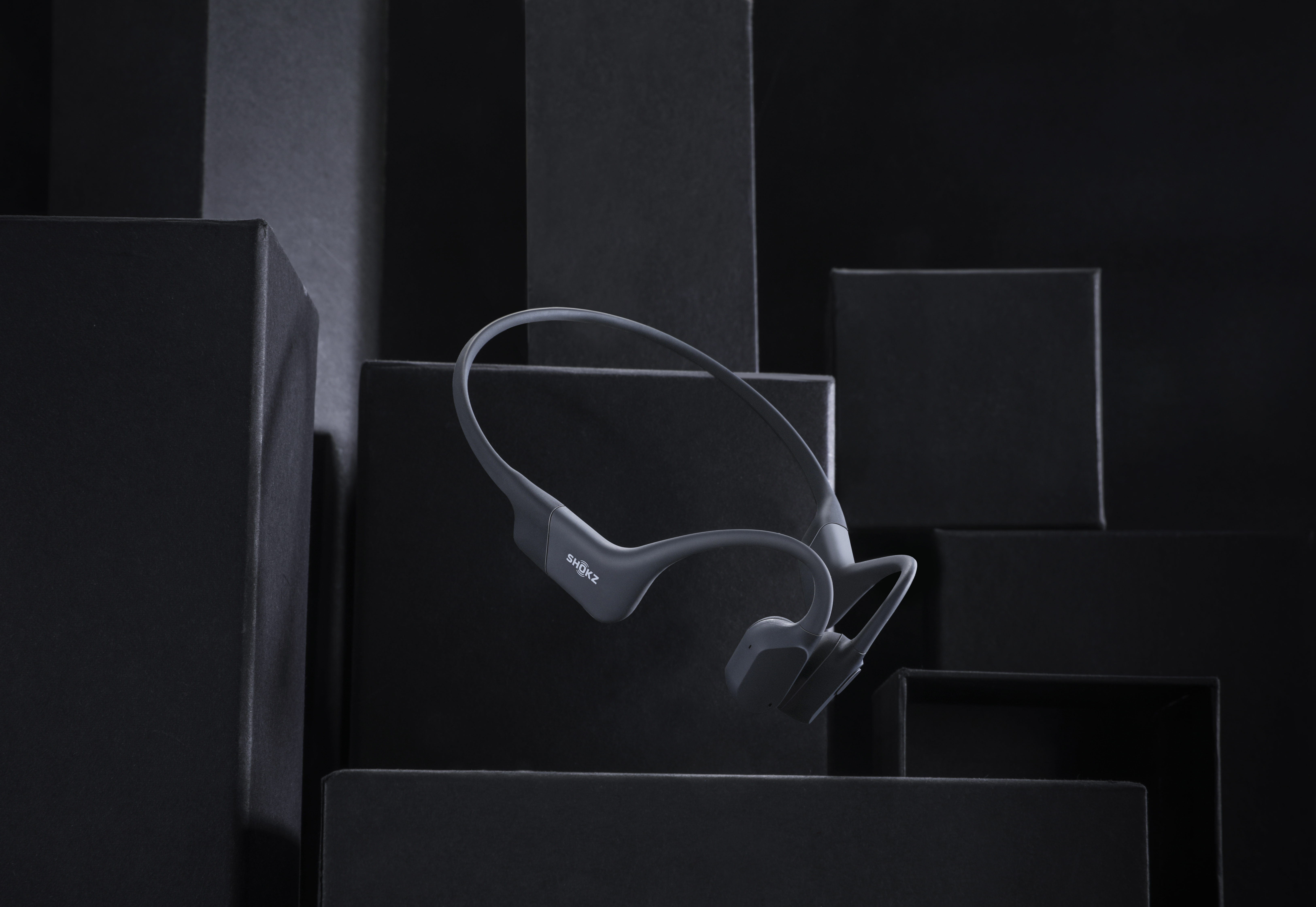 Szare słuchawki przewodnictwa kostnego. Umieszczone przed czarnymi kwadratowymi pudełkami.