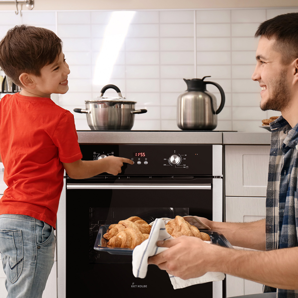 Ein Vater und Sohn in einer Küche. Der Sohn zeigt auf ein Bedienfeld des Backofens, während der Vater ein Blech mit Croissants hält.
