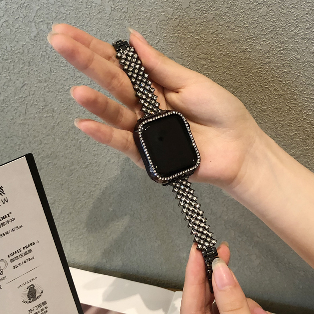 Hand hält eine Uhr mit schwarzem Bildschirm und einem geschmückten Band auf grauem Hintergrund.