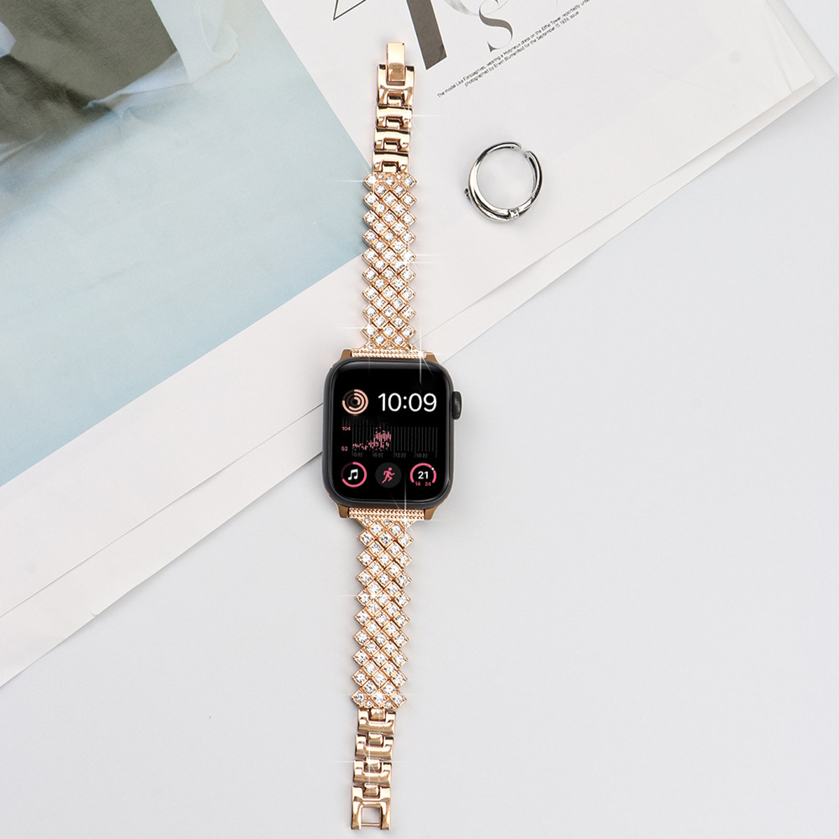 Roségoldene Smartwatch mit diamantähnlichem Band, zusammen mit einem Ring auf weißem Papier.