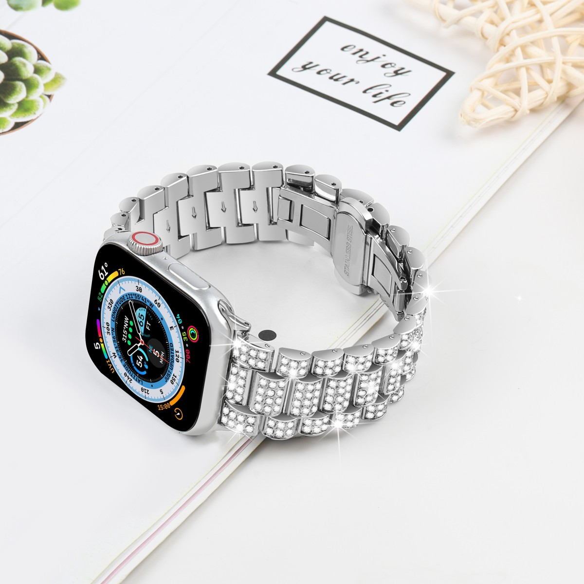 Silberne Apple Watch mit Schmuckband auf weißer Oberfläche. Papier mit Text 'enjoy your life' sichtbar.