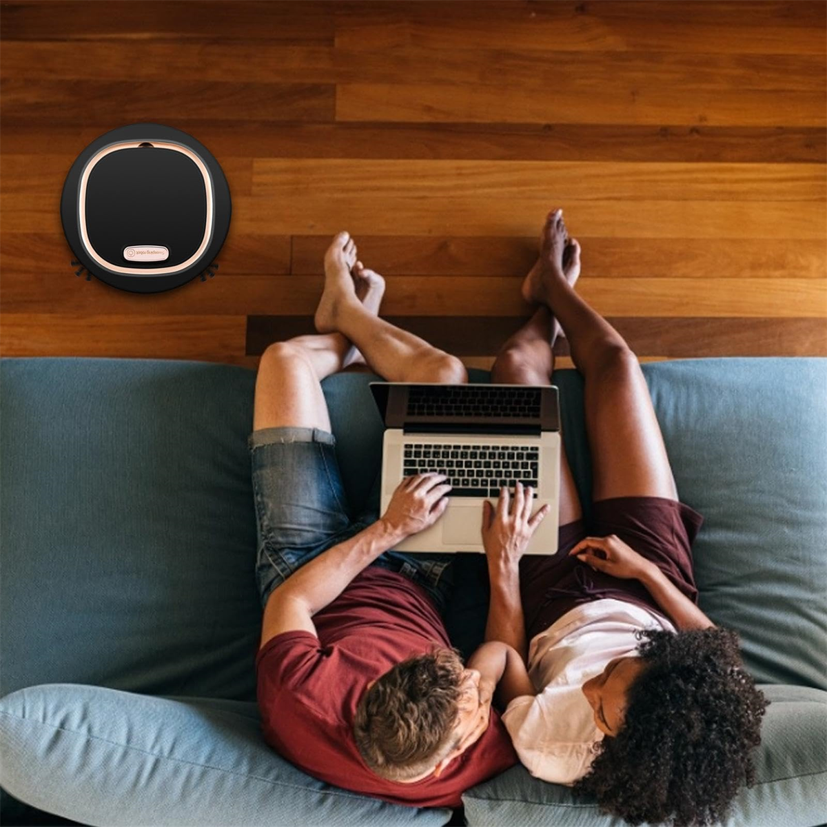 Paar auf Couch mit Laptop, ein Roboterstaubsauger in der Nähe.
