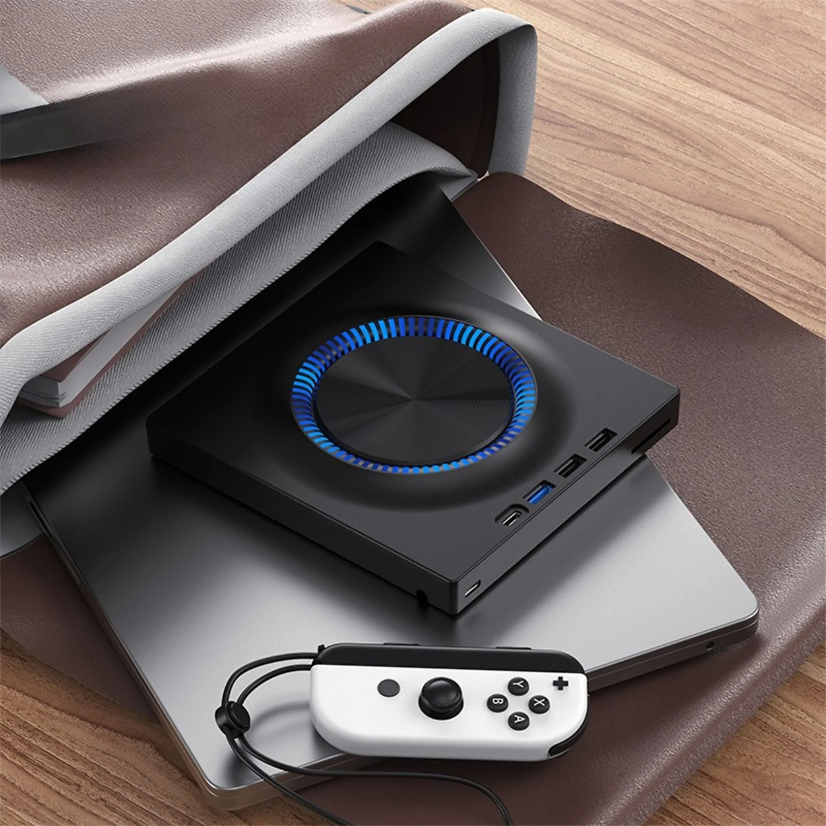 Schwarzes Gerät mit blauem Kreis und Anschlüssen, in einer Tasche auf einem Laptop. Ein weißer Controller daneben.