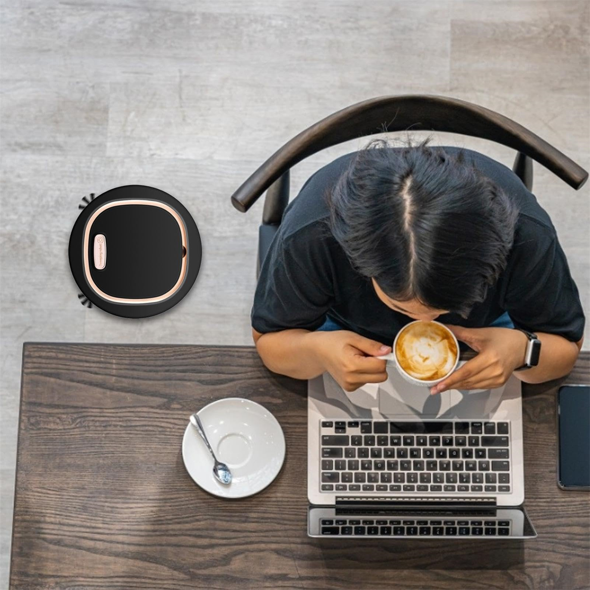 Person trinkt Kaffee, mit Laptop und Roboterstaubsauger auf Holztisch.