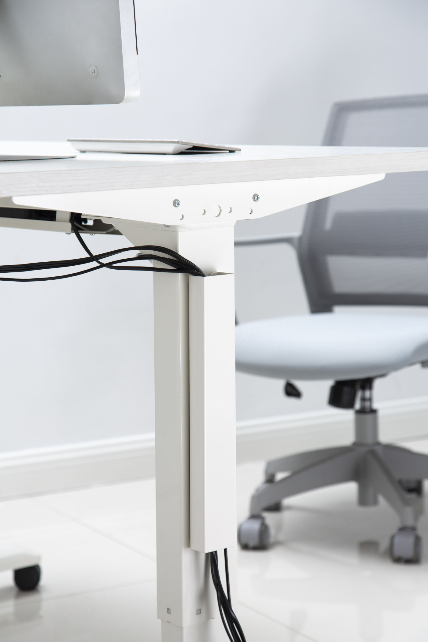 Ein weißer Schreibtisch mit einem Monitor und einem grauen Bürostuhl. Kabel verlaufen unter dem Schreibtisch.