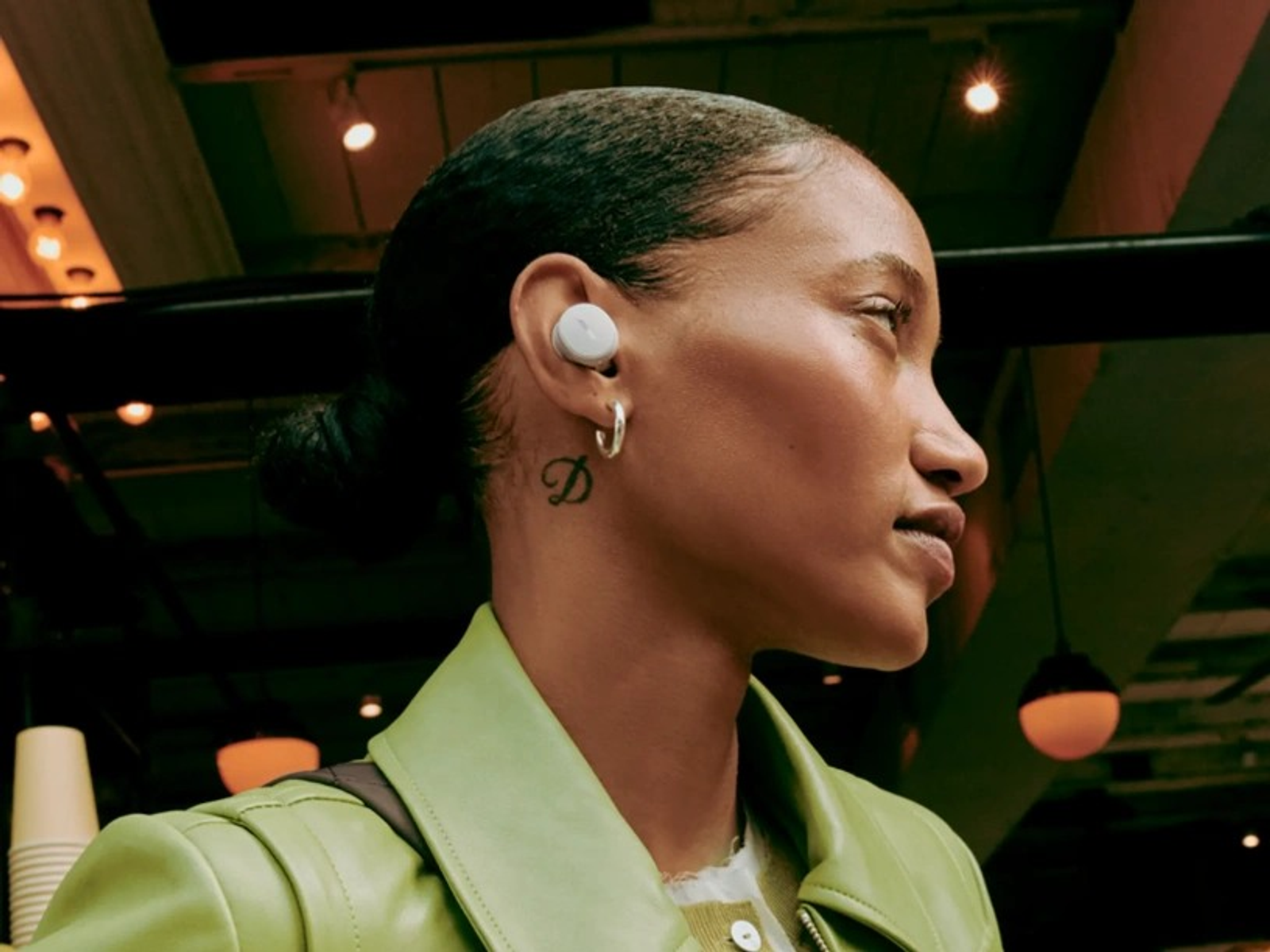 Mujer con auriculares blancos. Vista de perfil, pelo recogido, chaqueta verde. Fondo borroso.