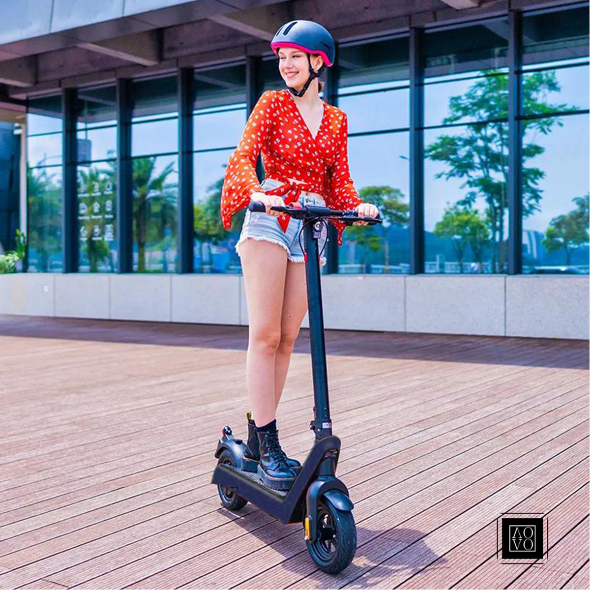 Młoda kobieta na hulajnodze elektrycznej, w kasku i czerwonej górze, na drewnianym chodniku.