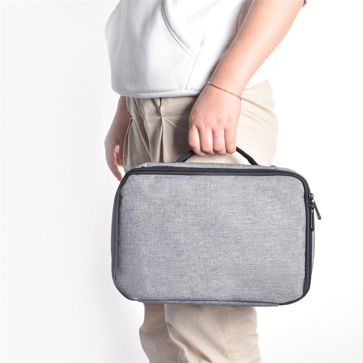 Eine Person hält eine graue rechteckige Tasche mit Reißverschluss. Die Person trägt einen weißen Pullover.