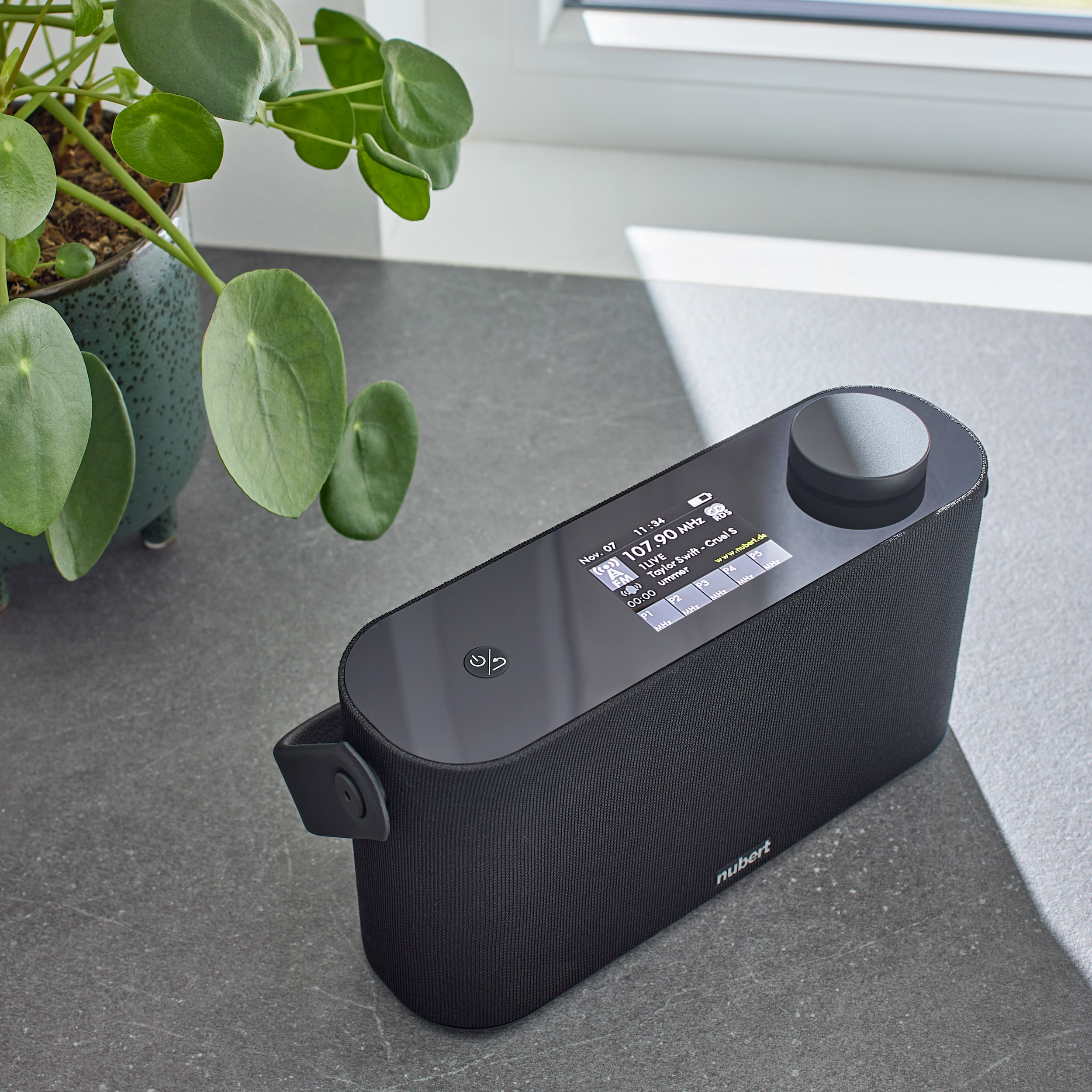 Schwarzes und silbernes Audiogerät. Es steht auf einer Theke neben einer Pflanze. Zeigt ein Display.