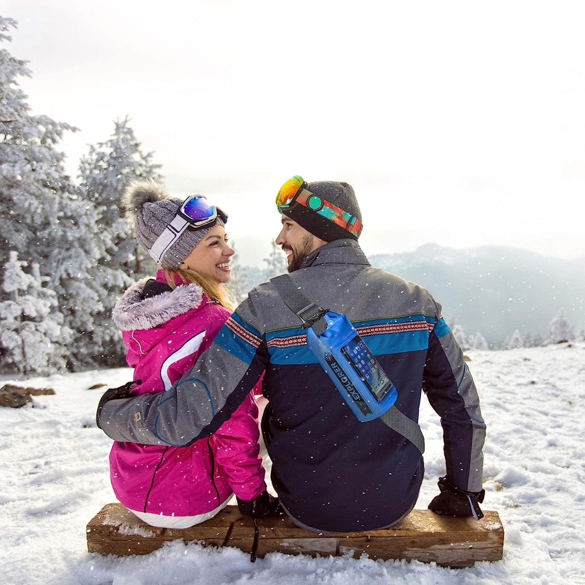 Una pareja sentada en un banco de madera, en montañas nevadas, con equipo de esquí, gafas y botella.