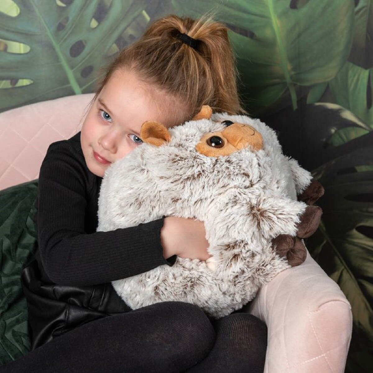 Ein Mädchen umarmt ein flauschiges Schafspielzeug, während es sitzt. Sie hat einen Pferdeschwanz und trägt ein schwarzes Outfit.