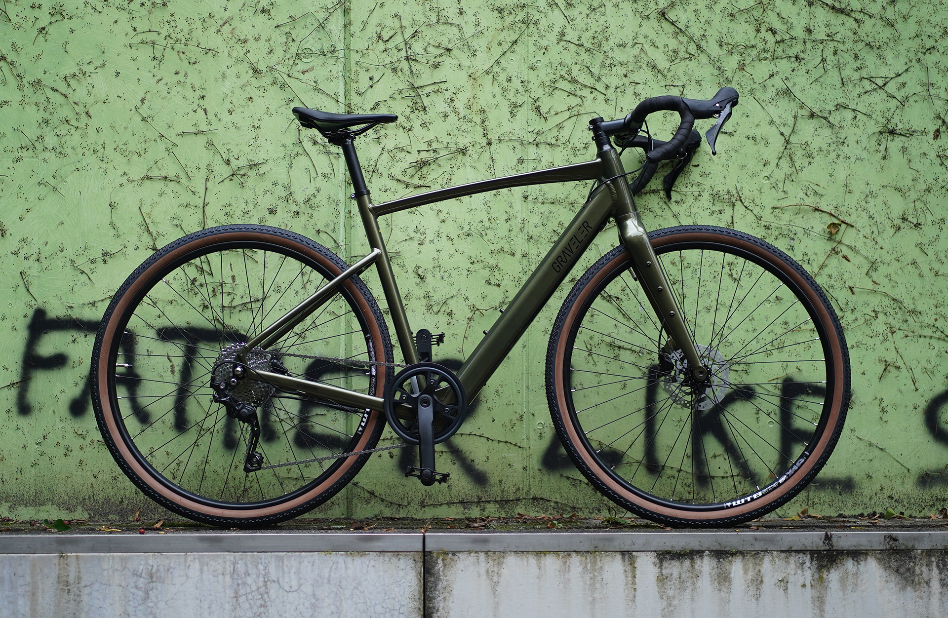 Dunkelgrünes Gravelbike mit braunen Reifen vor grünem Graffiti.