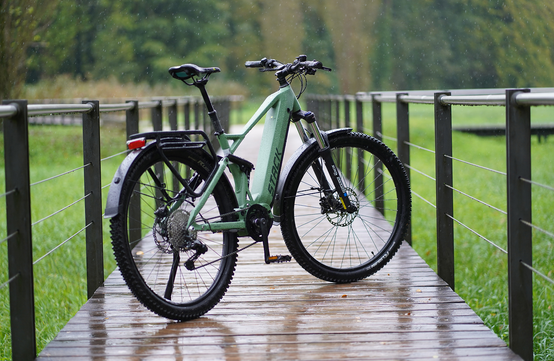Ein grünes E-Bike steht im Regen auf einer Holzbrücke. Der Hintergrund ist ein grüner Wald.