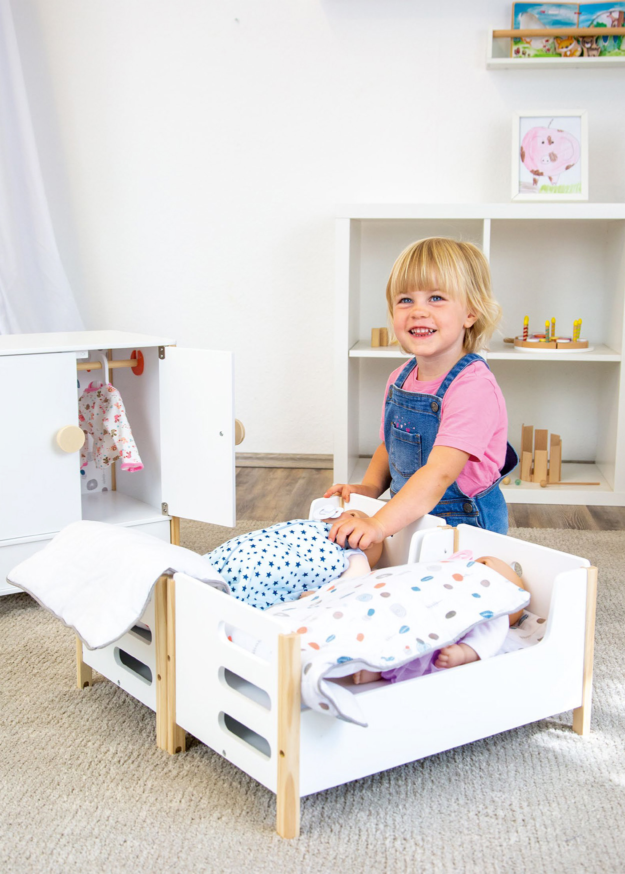 Junges Kind spielt mit Puppen in einem weißen Bett, lächelt. Kleiderschrank und Regale im Hintergrund.