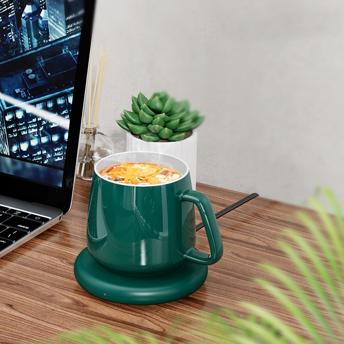 Grüner Becher auf einer Heizplatte. Pflanze und Laptop stehen auf dem Holztisch.