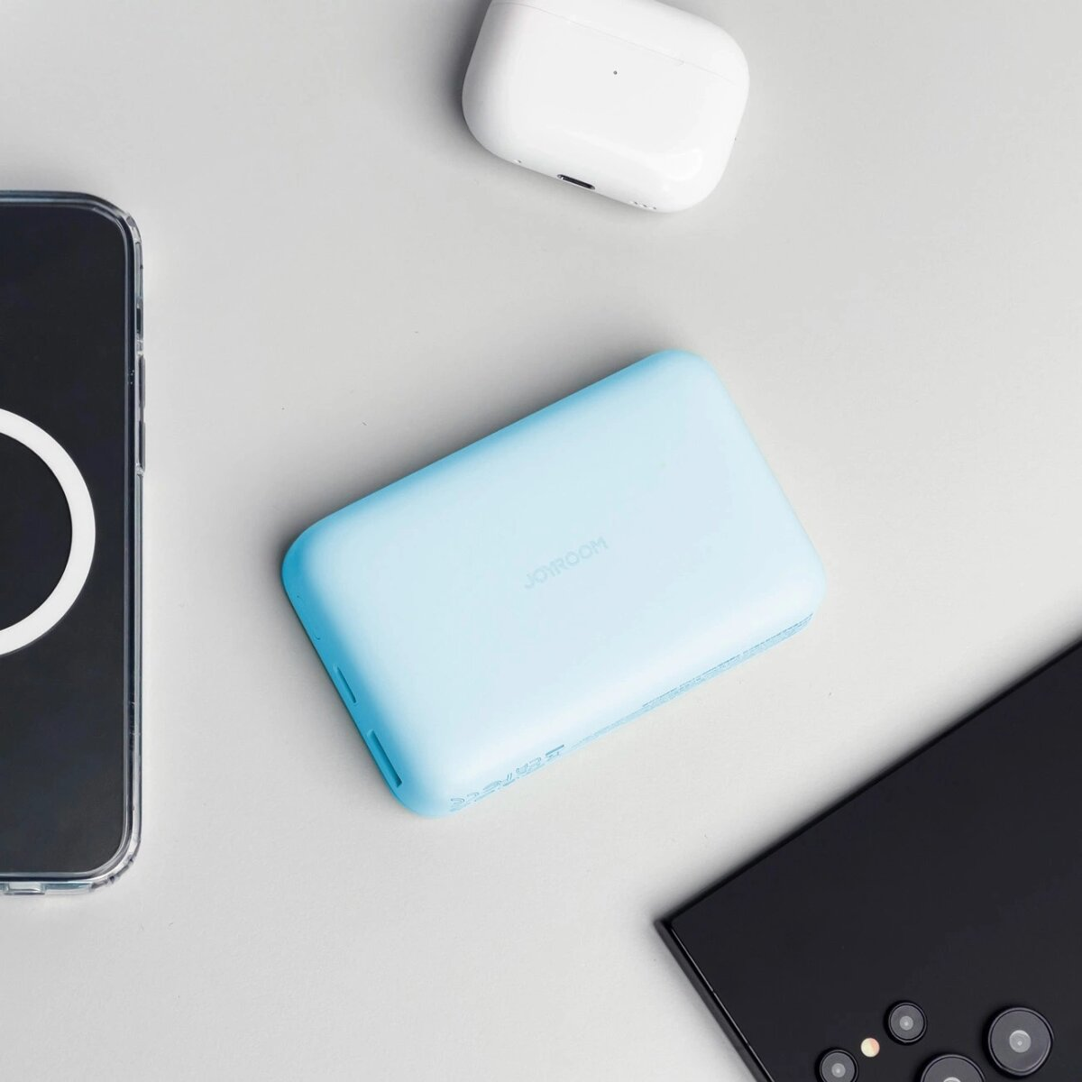 Eine hellblaue Powerbank, ein Telefon, ein weißes Ohrhörer-Etui und ein schwarzes Telefon liegen auf einer Oberfläche.