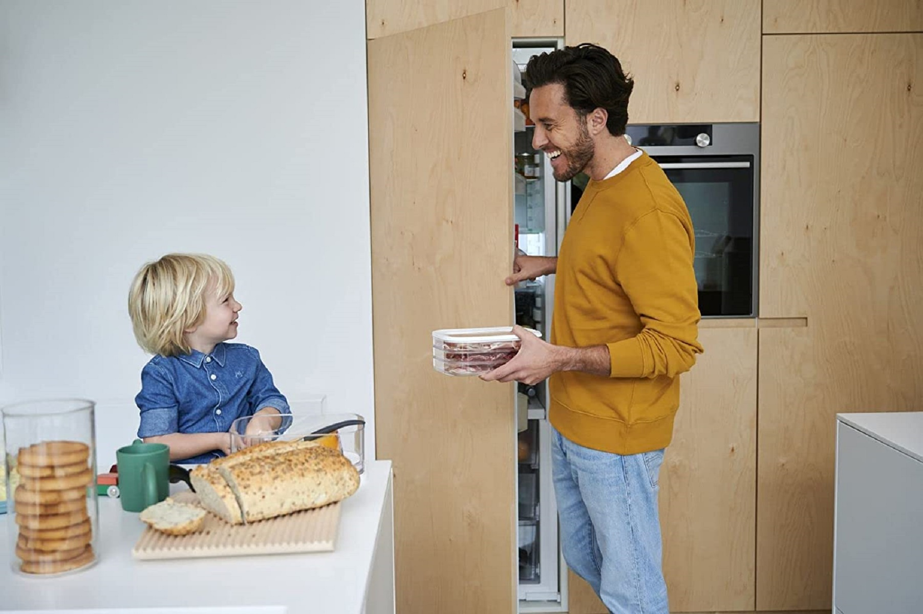 Ein Mann und ein Kind in einer Küche. Der Mann öffnet einen Kühlschrank, das Kind sitzt am Tisch.