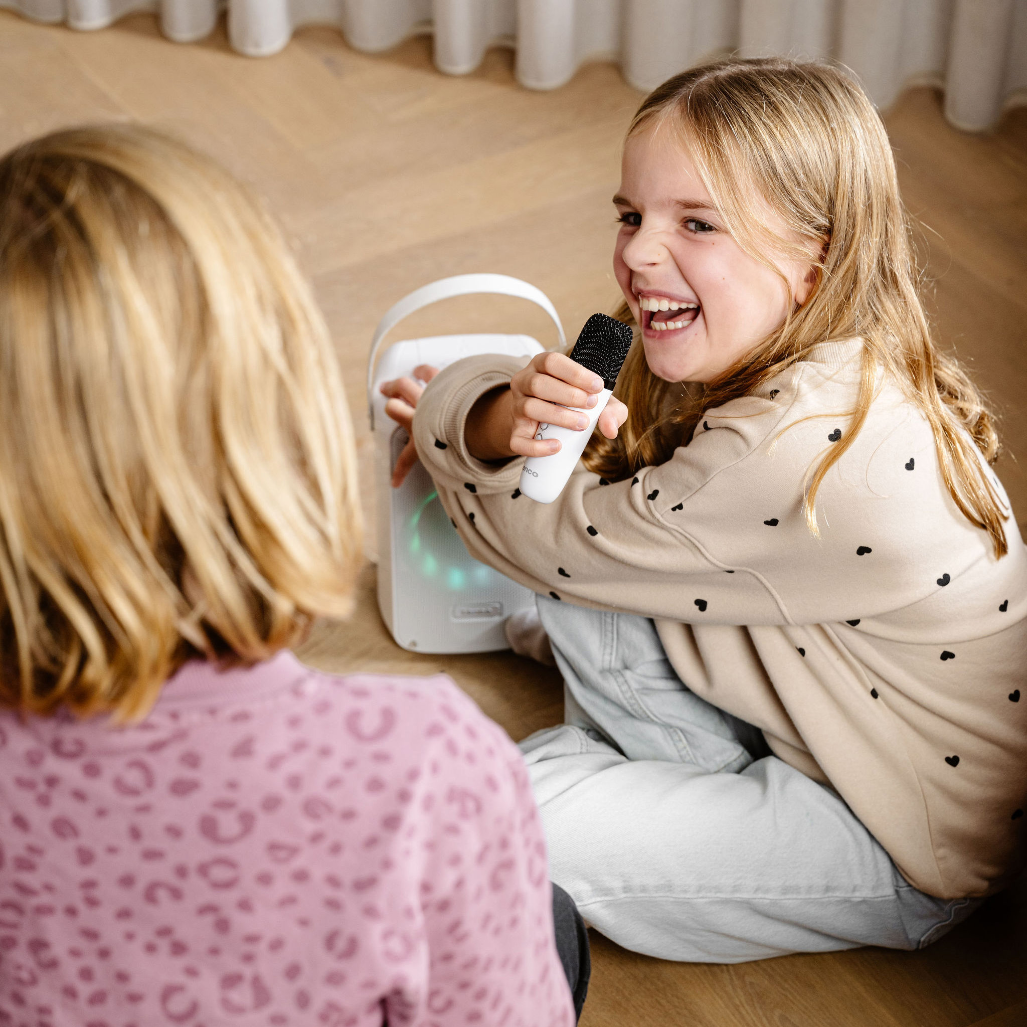 Twee jonge meisjes zingen in een microfoon die is aangesloten op een luidspreker. Eén meisje glimlacht breed.