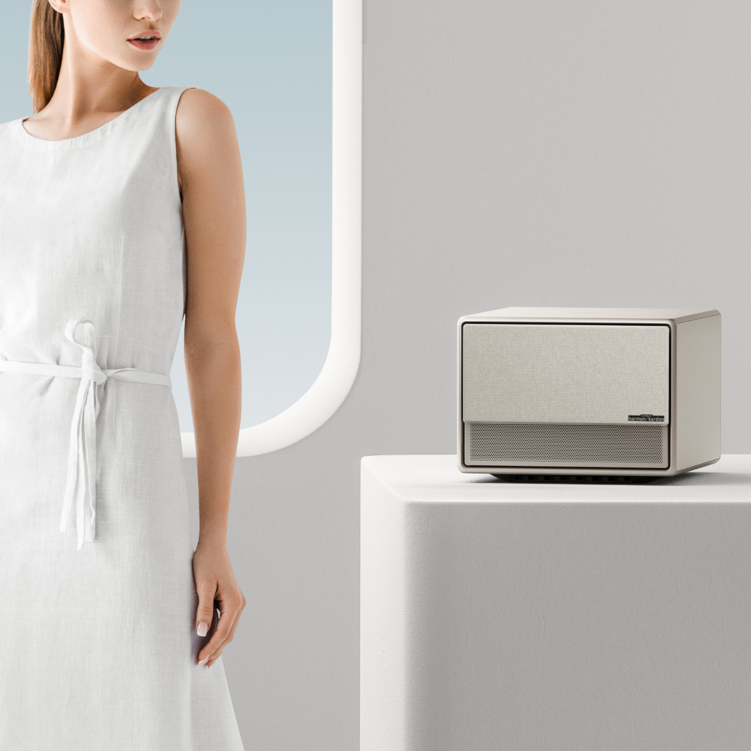 Kobieta w białej sukience obok głośnika na cokole. Neutralne tło.