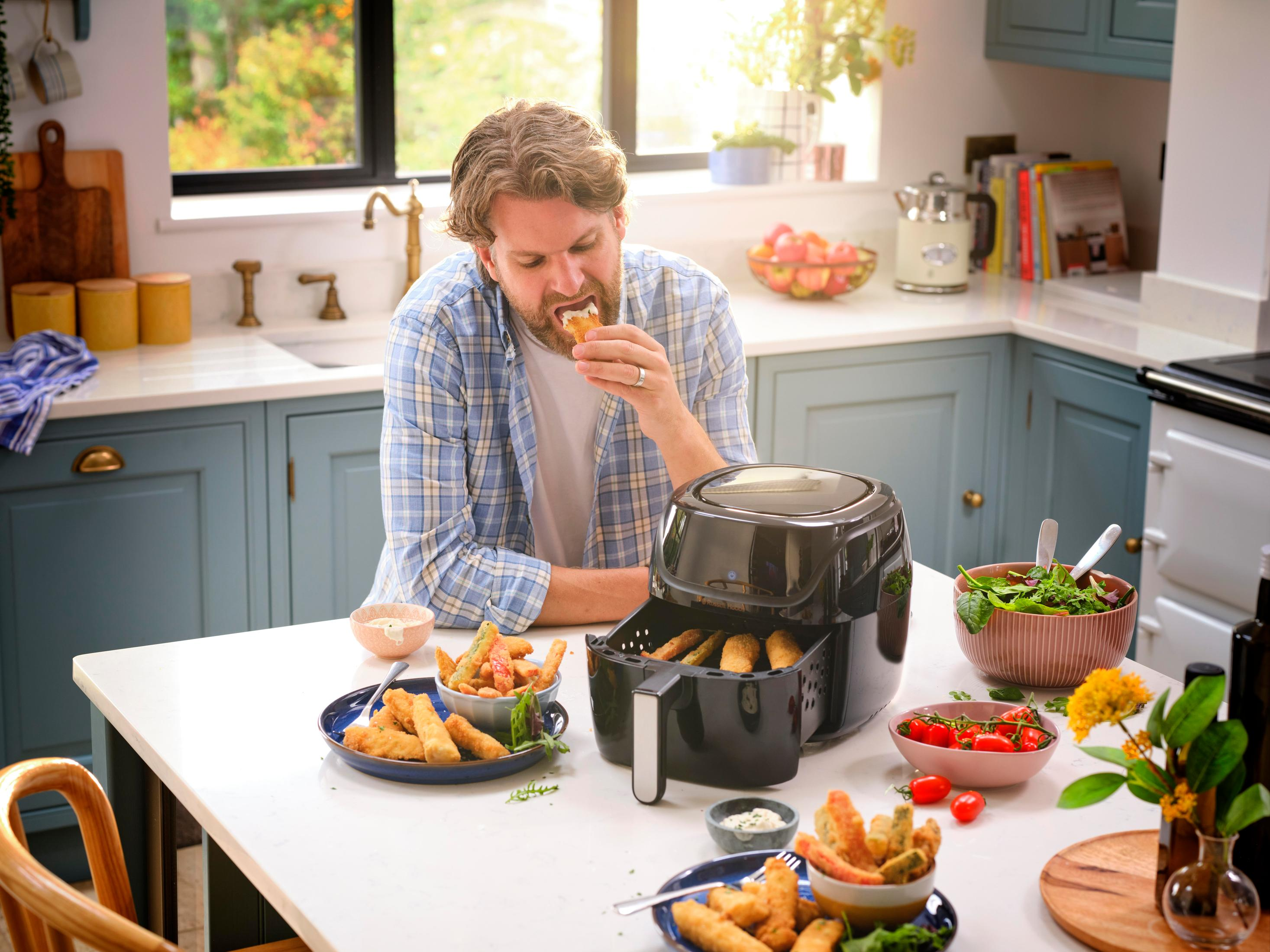 Uomo mangia cibo fritto da una friggitrice ad aria nera. Cucina con altri cibi.