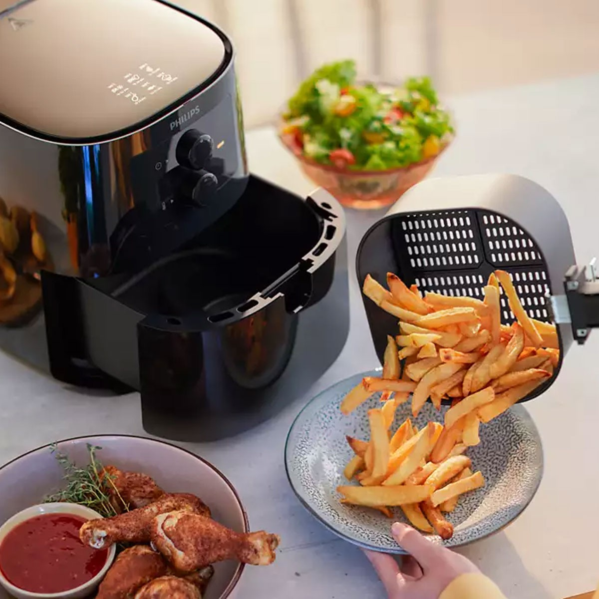 Freidora de aire con papas fritas vertiéndose en un bol. Pollo y ensalada presentes.