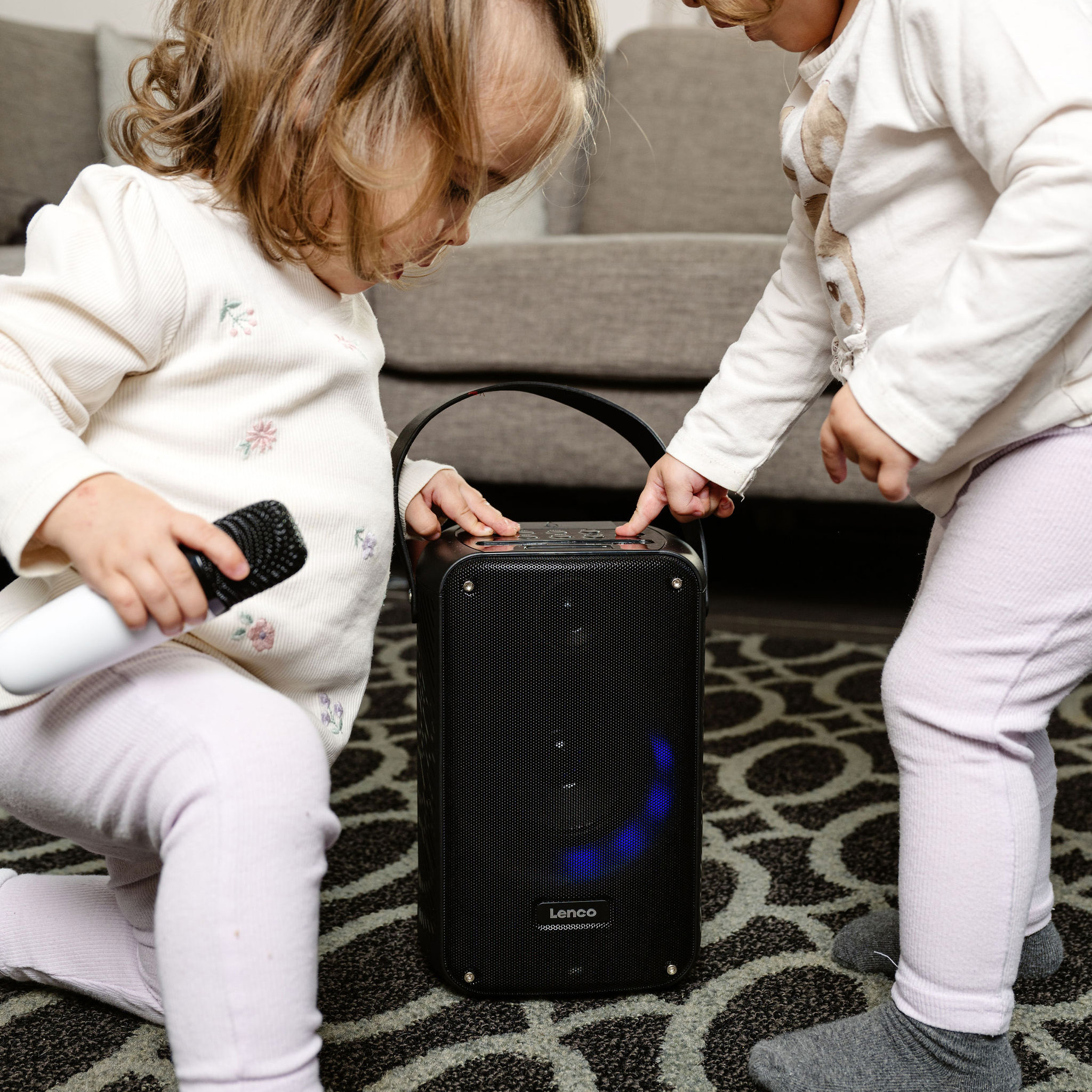 Twee jonge kinderen spelen met een zwarte luidspreker. Eén houdt een microfoon vast. Een kleed met patroon op de achtergrond.