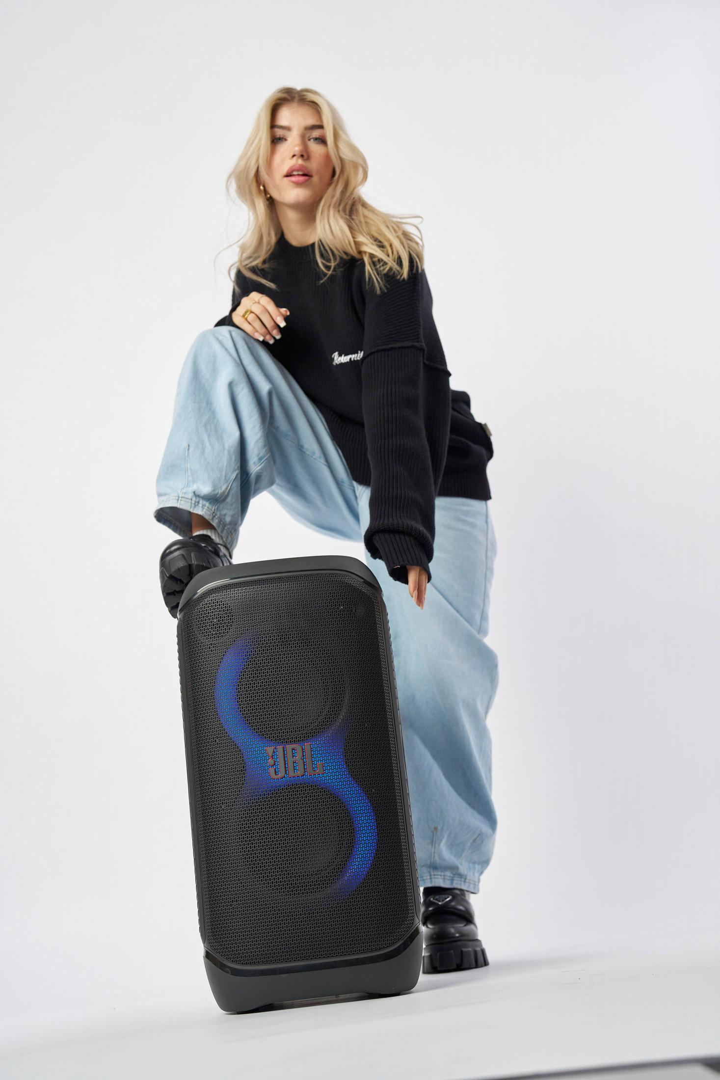 Eine Frau in einem schwarzen Pullover und hellblauen Jeans steht neben einem schwarzen JBL-Lautsprecher. Weißer Hintergrund.
