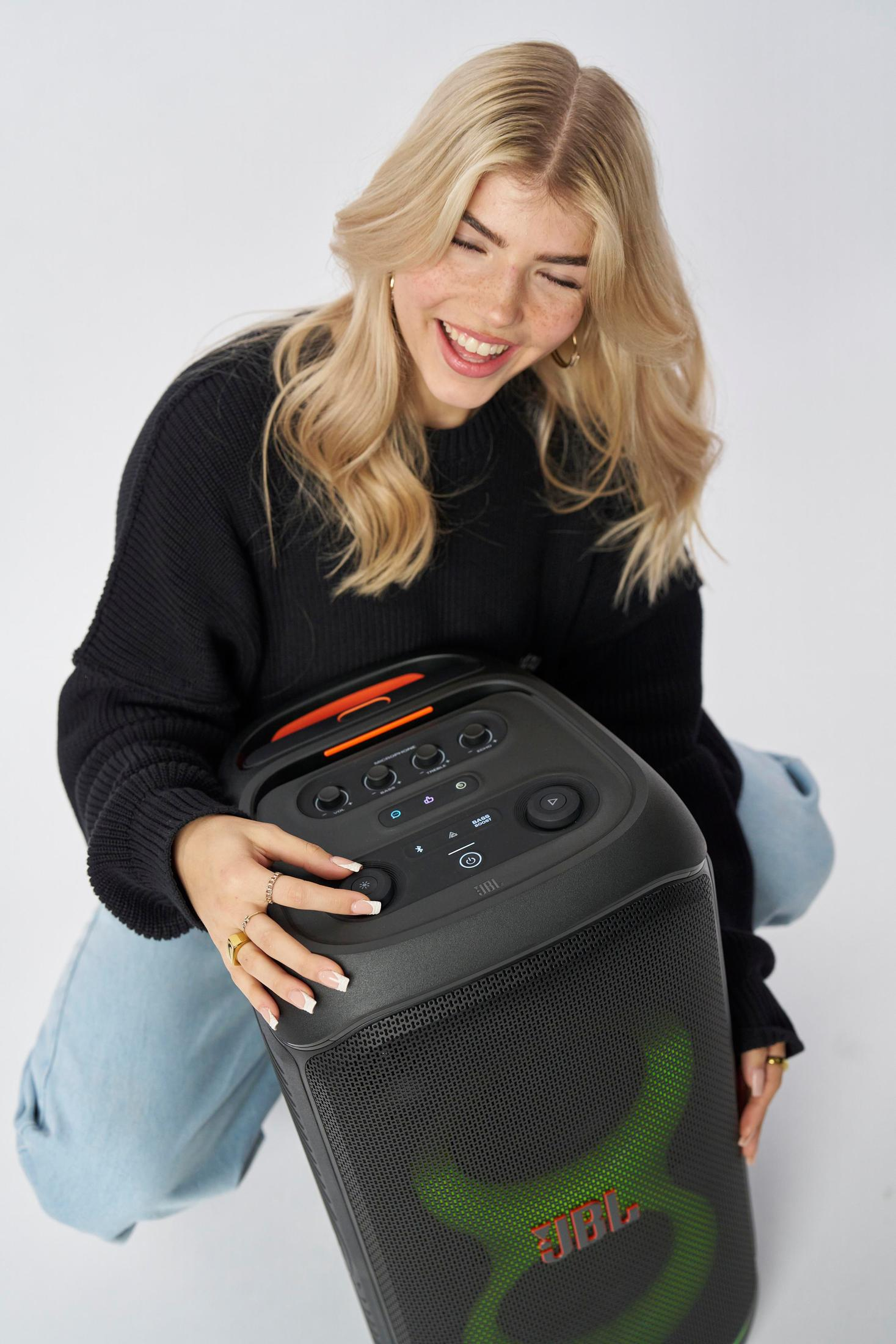 Blonde Frau lächelt, hält schwarzen JBL-Lautsprecher. Weißer Hintergrund.