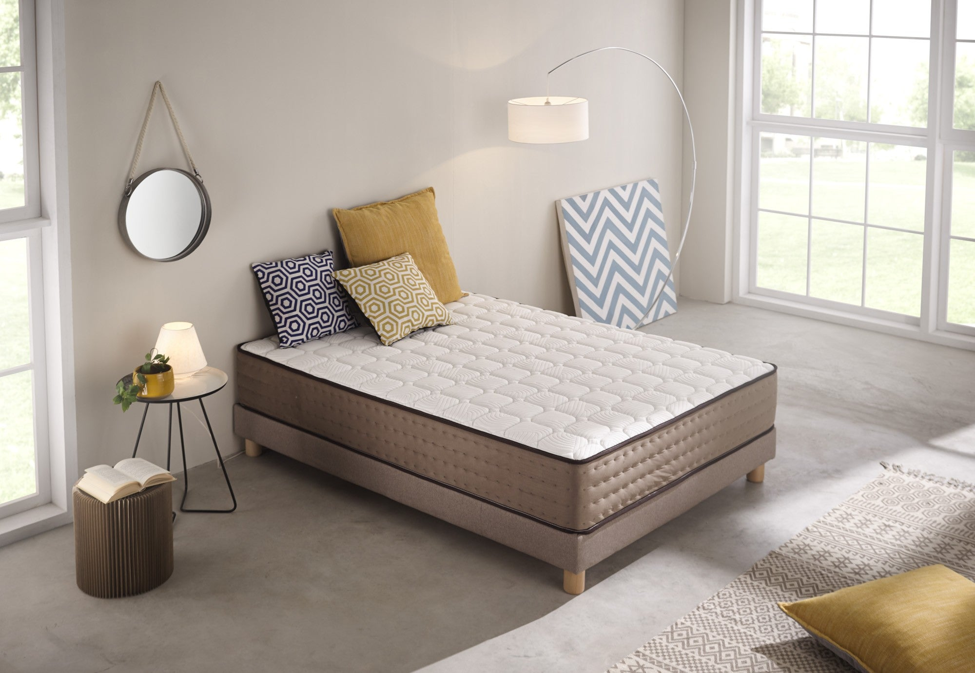 Una cama en una habitación. El colchón es blanco con borde marrón. También hay almohadas, una lámpara, un espejo y una alfombra estampada.