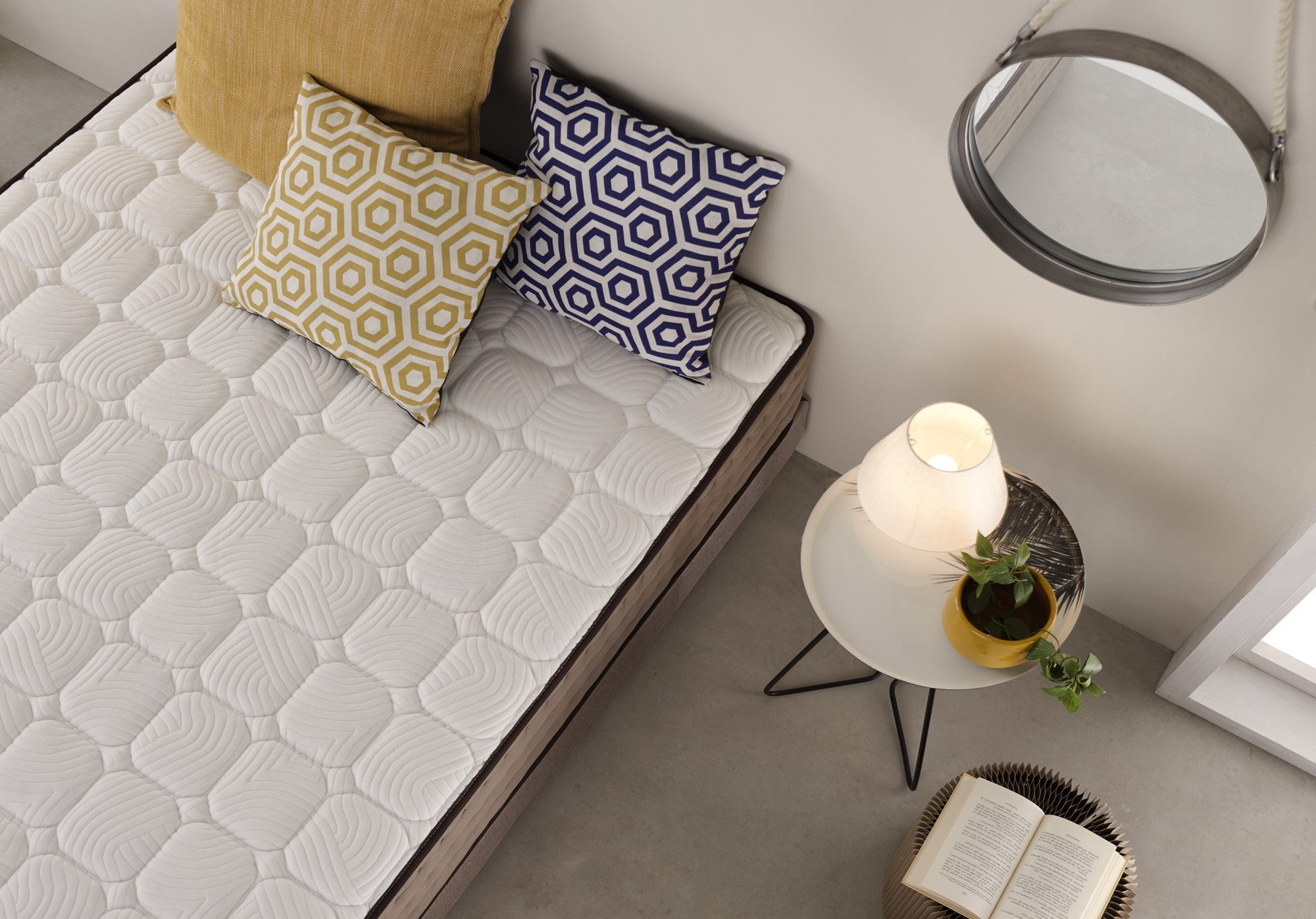 Cama con colchón, almohadas y mesa auxiliar con lámpara y libro. Espejo en la pared.