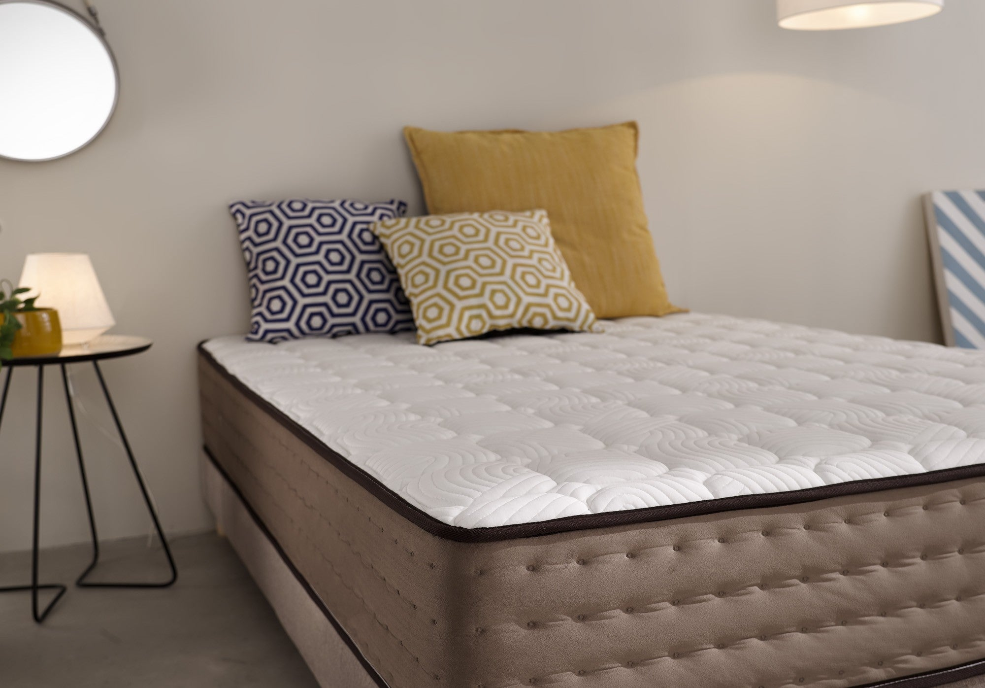 Cama con colchón, almohadas y mesa auxiliar con lámpara. Espejo en la pared.