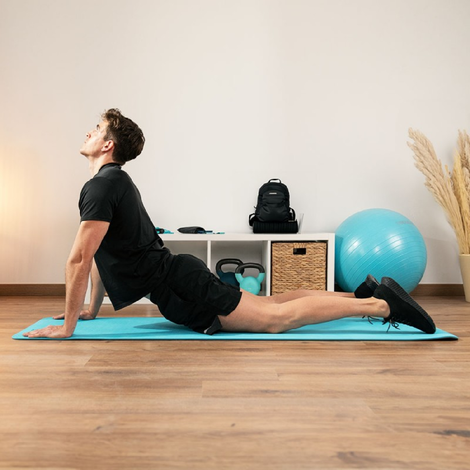 Uomo su un tappetino blu che fa una posa yoga, con una palla e altri oggetti sullo sfondo.