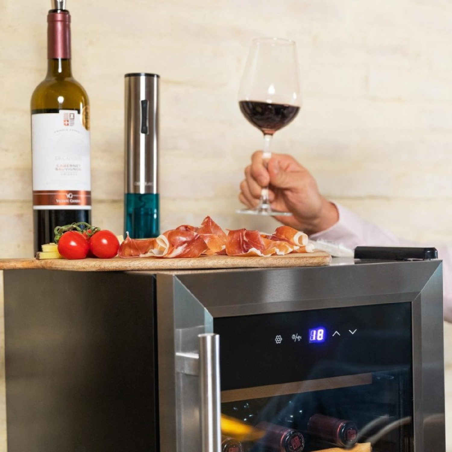 Persona sosteniendo una copa de vino tinto, junto a una vinoteca, comida y sacacorchos.