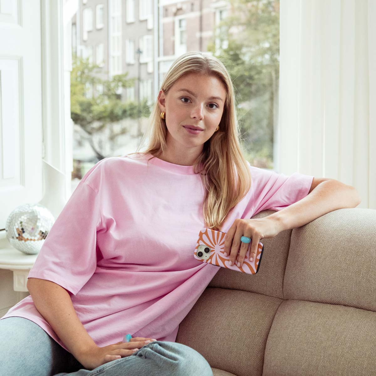Een vrouw in een roze shirt zit op een beige bank en houdt een telefoonhoesje vast. Ze heeft gouden hoepels en een blauwe ring.