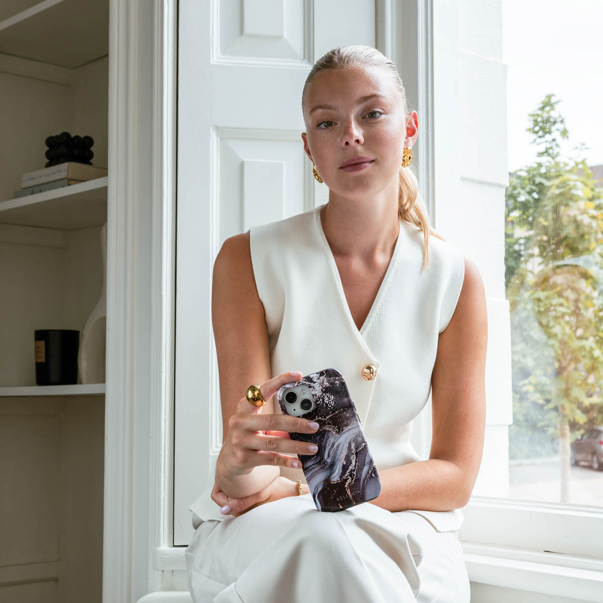 Een vrouw met blond haar houdt een telefoon met een marmeren hoesje vast. Ze draagt oorbellen.