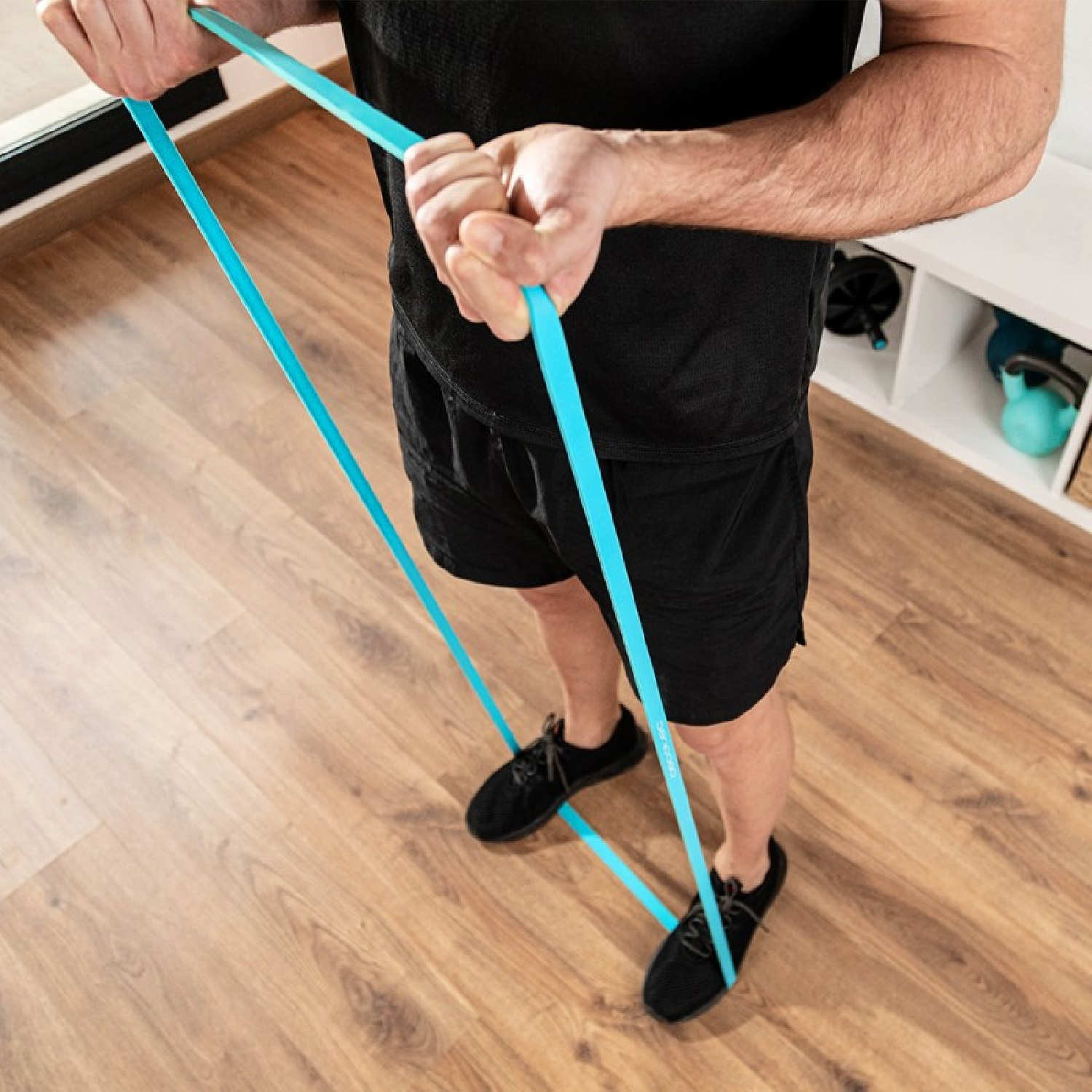 Hombre sujetando una banda elástica azul, estirándola. Pantalones cortos y zapatos negros, suelo de madera.