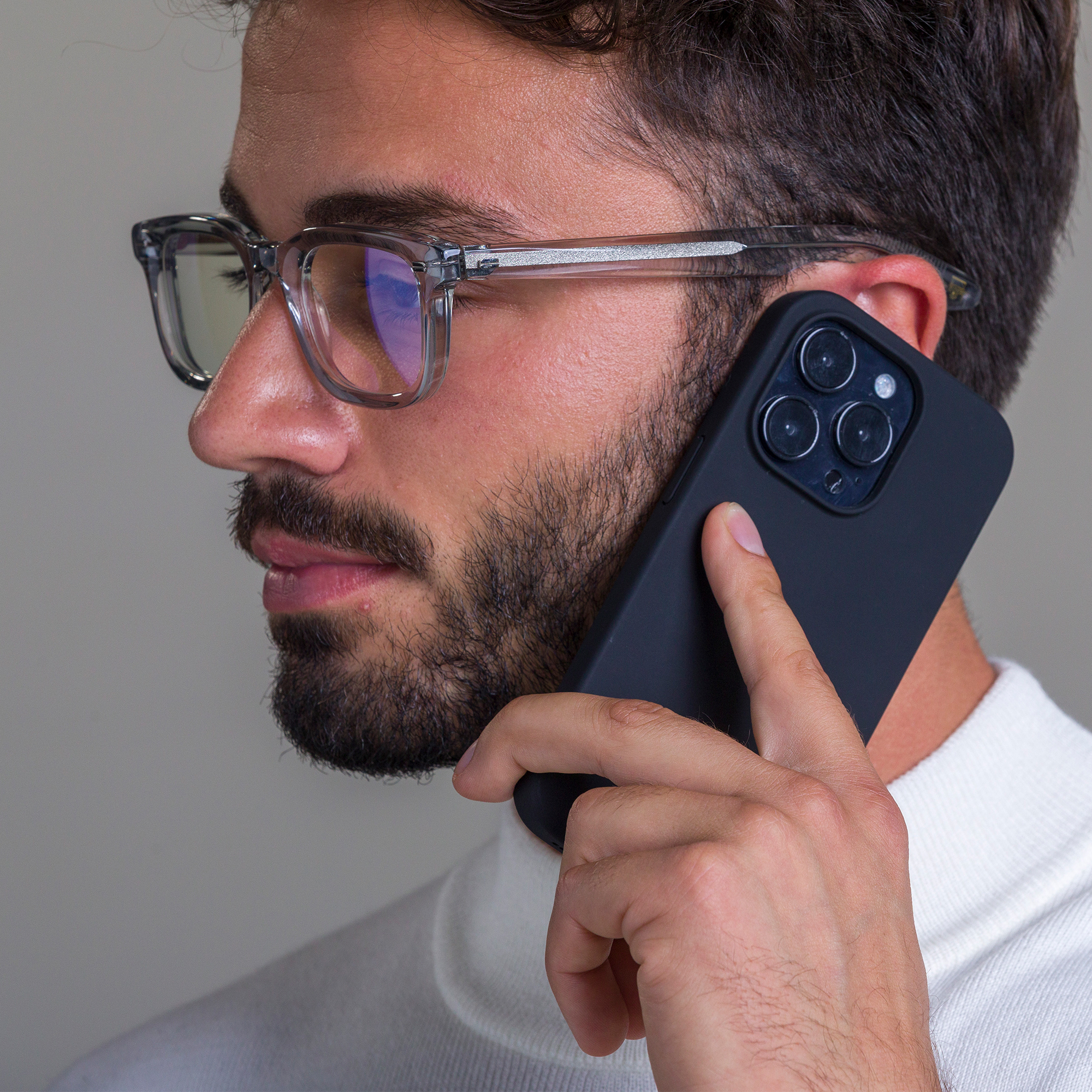 Uomo con occhiali e barba tiene telefono nero, con maglione bianco. Profilo.