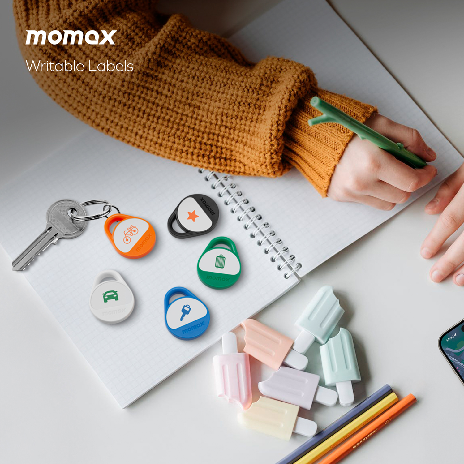 A desk with momax labels, key, pens and cell phone. A person is writing with a pen.
