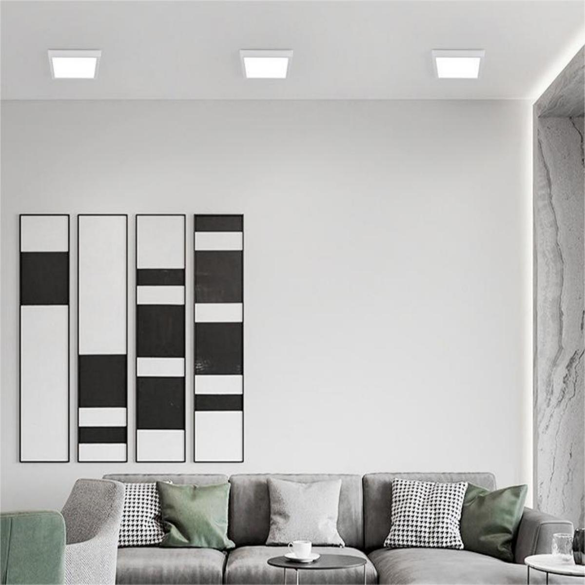 Drei quadratische LED-Deckenleuchten in einem weißen Raum, mit abstrakter Kunst und einem Sofa.
