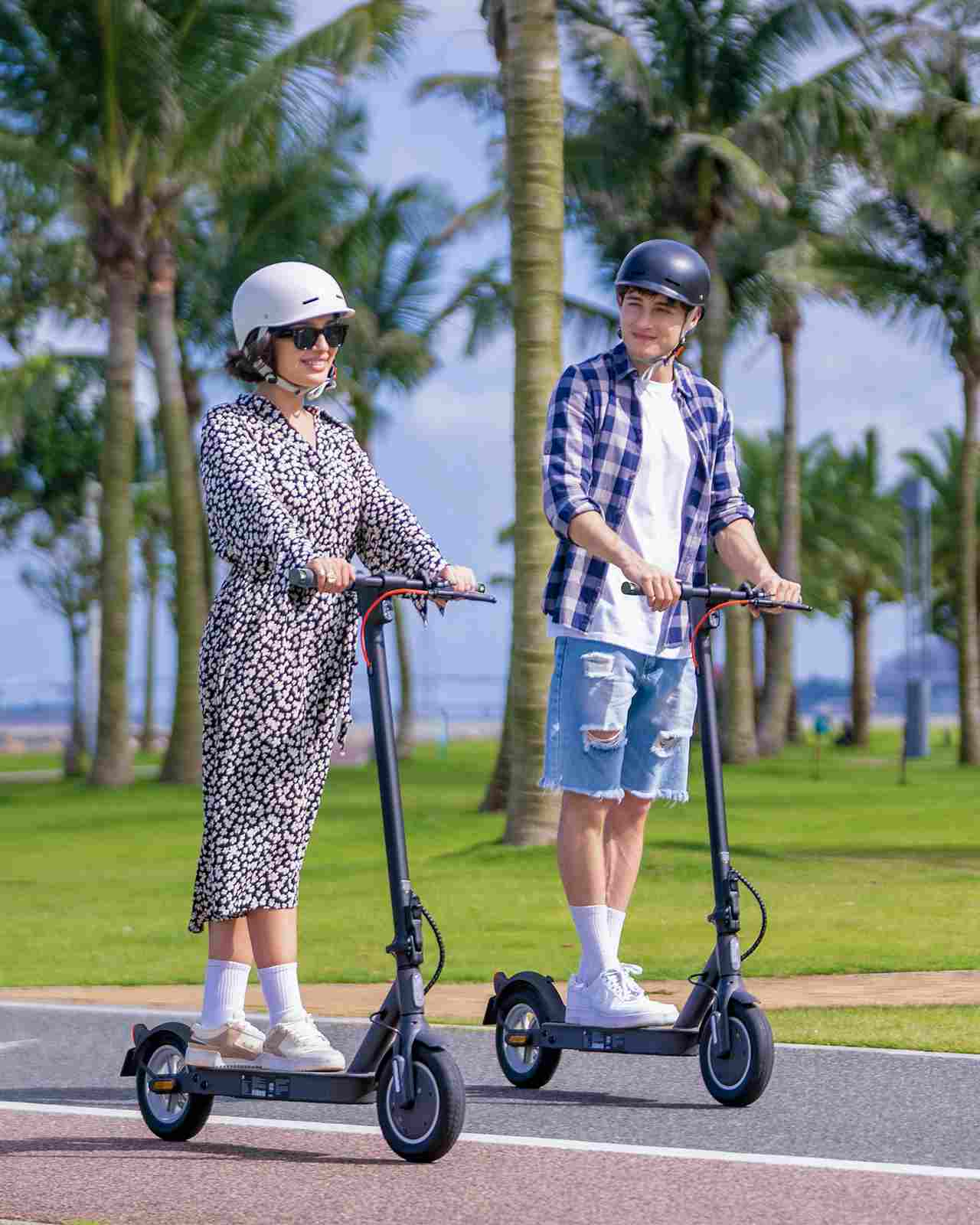 Zwei Personen fahren mit Elektrorollern auf einem asphaltierten Weg. Palmen im Hintergrund.