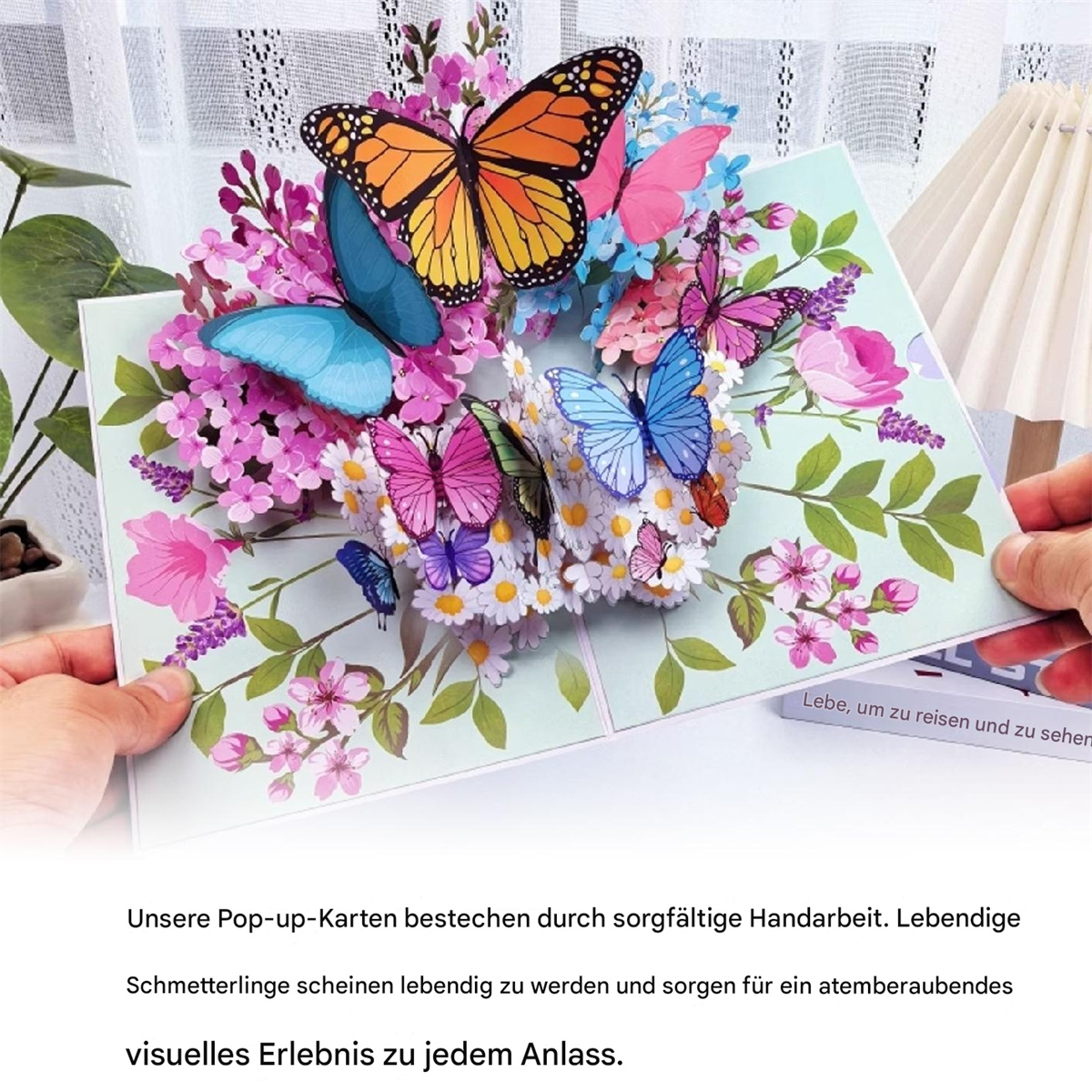 Pop-up-Karte mit bunten Schmetterlingen und Blumen. Hände halten sie offen. Hintergrund ist ein Fenster mit Vorhängen.