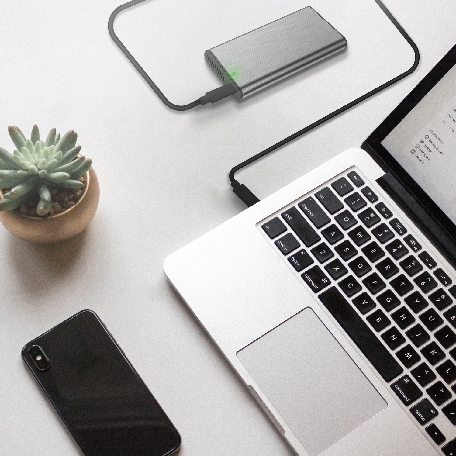 Laptop, externe harde schijf, telefoon en kleine plant op een wit oppervlak. Stroomkabel is aangesloten.