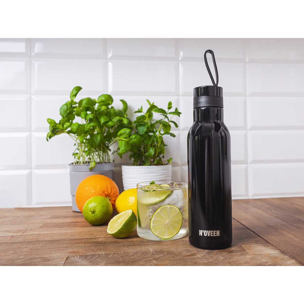 Eine schwarze Wasserflasche mit Griff. Sie steht auf einem Holztisch mit Obst, Pflanzen und einem Glas.