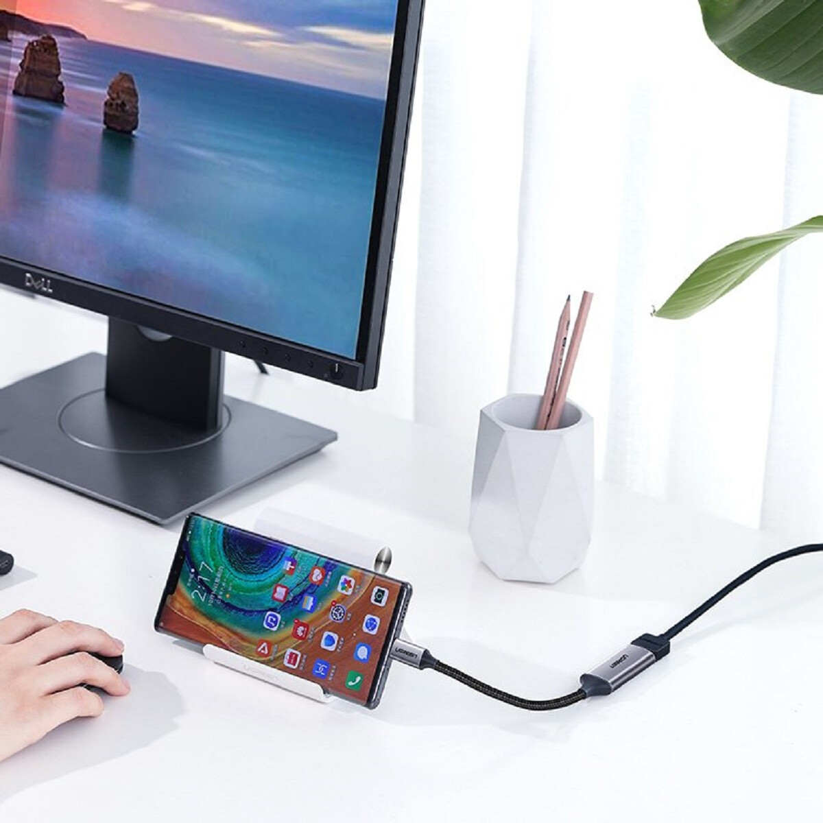 Eine Person, die einen Computer benutzt, mit einem Telefon, das an einen Monitor angeschlossen ist. Ein Stifthalter steht auf dem Schreibtisch.